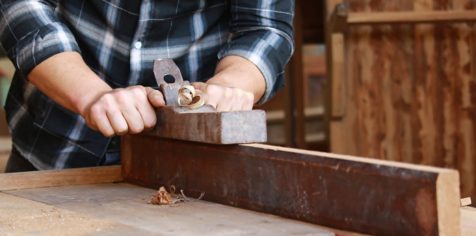 Carpenter using a hand plane to smooth a wood beam, with curled shavings on the workbench
